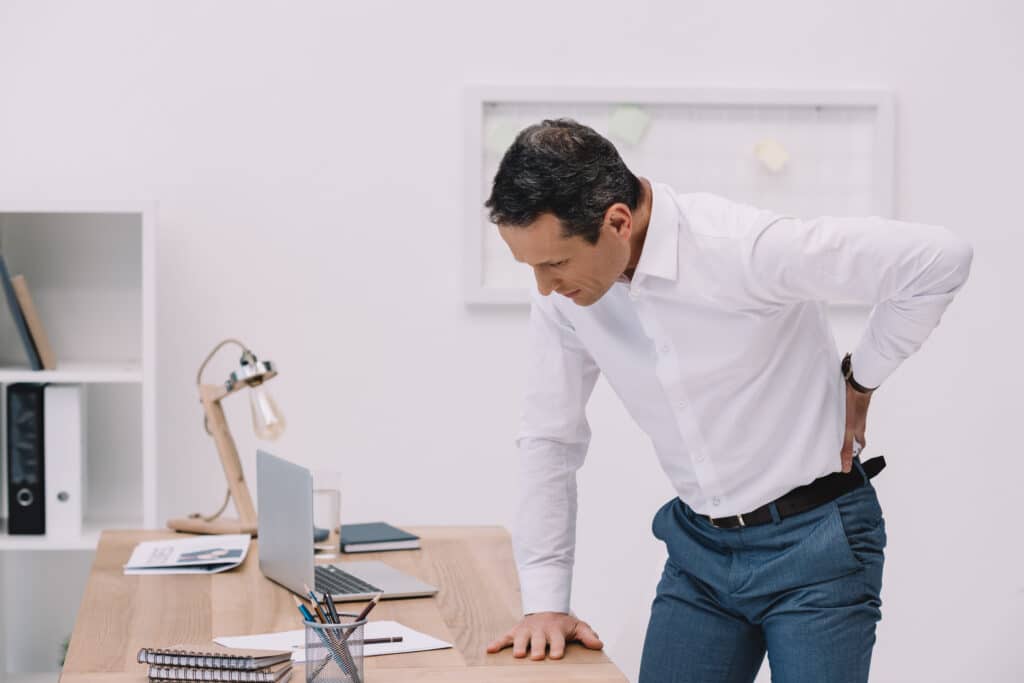 A man stands at his desk holding his tight back